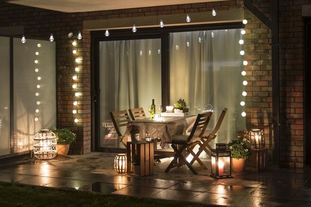 A backyard patio dining set at night, surrounded by string lights, lanterns, and potted flowers.